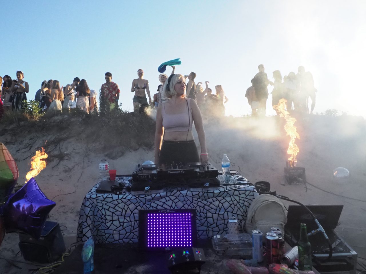 Daniela DJing on the beach at sunrise, flames either side and dancers along the dune