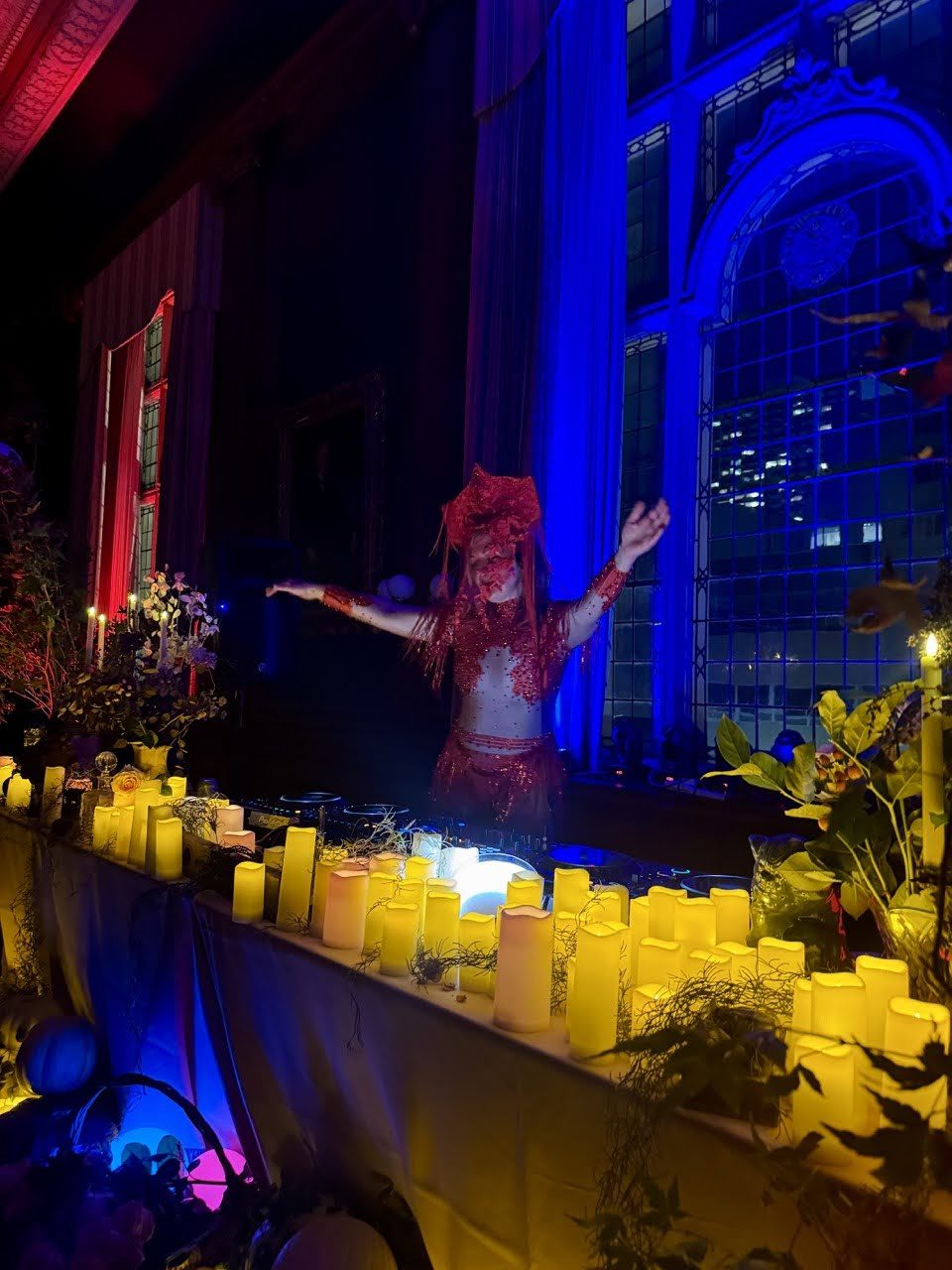 Daniela in red with arms outstretched behind rows of candles, a blue-lit arched window behind her
