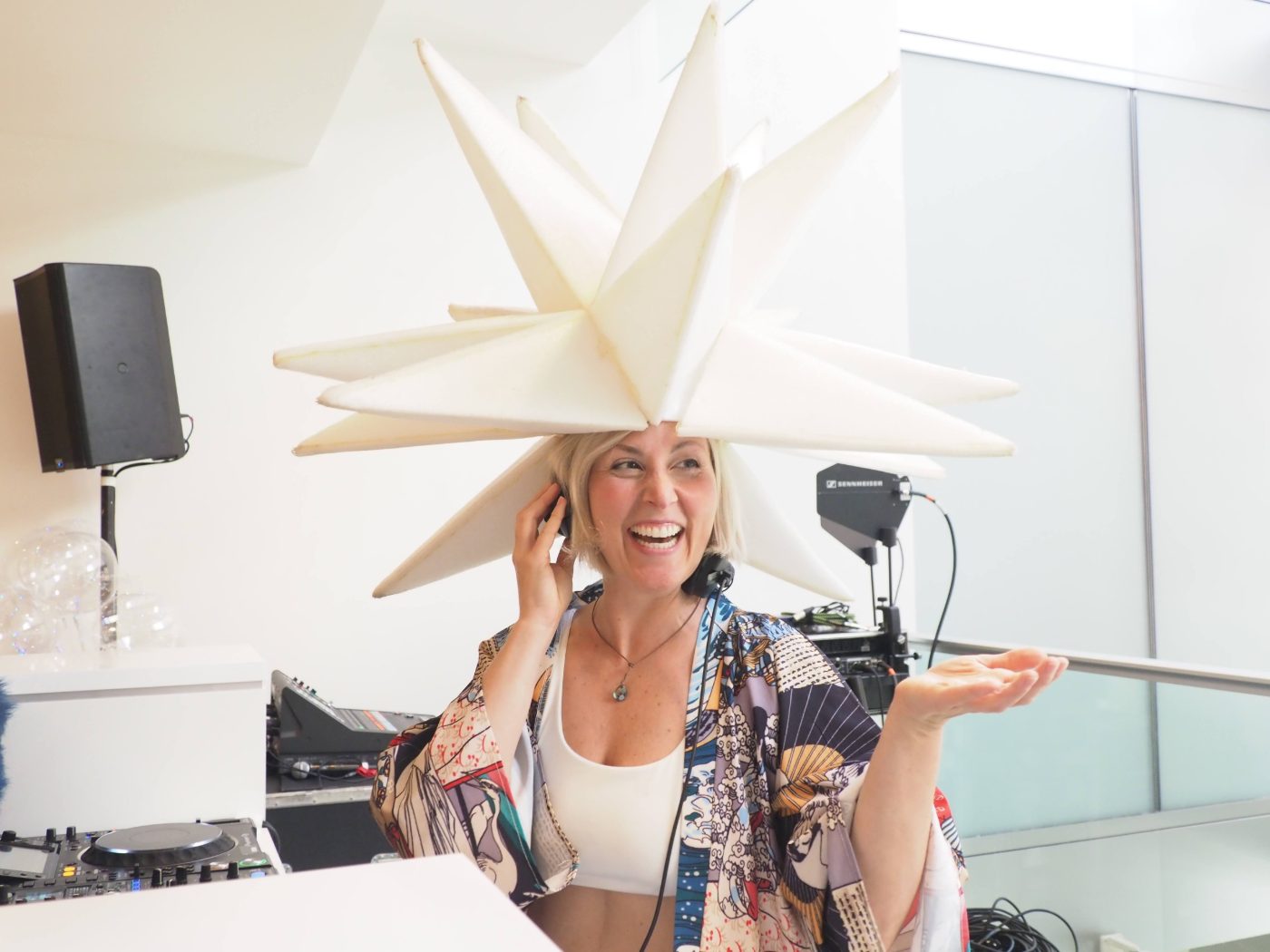 Daniela laughing behind the decks in a giant white star headpiece, one hand to her headphones