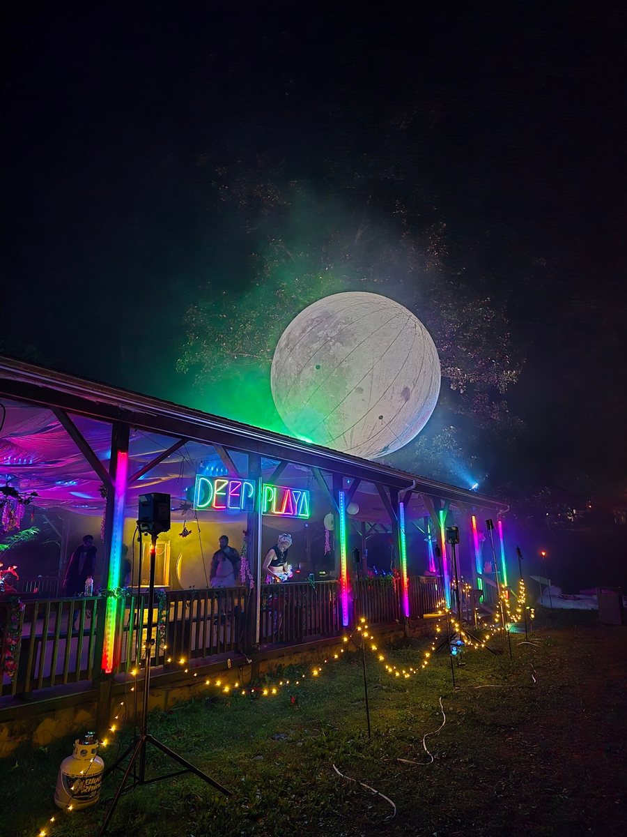 The Pavilion at night from outside: a giant glowing moon above the roof, a neon Deep Playa sign and rainbow LED tubes