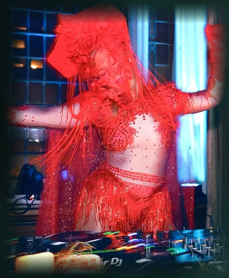 Daniela in a red beaded headdress and fringed costume, arms raised behind the decks